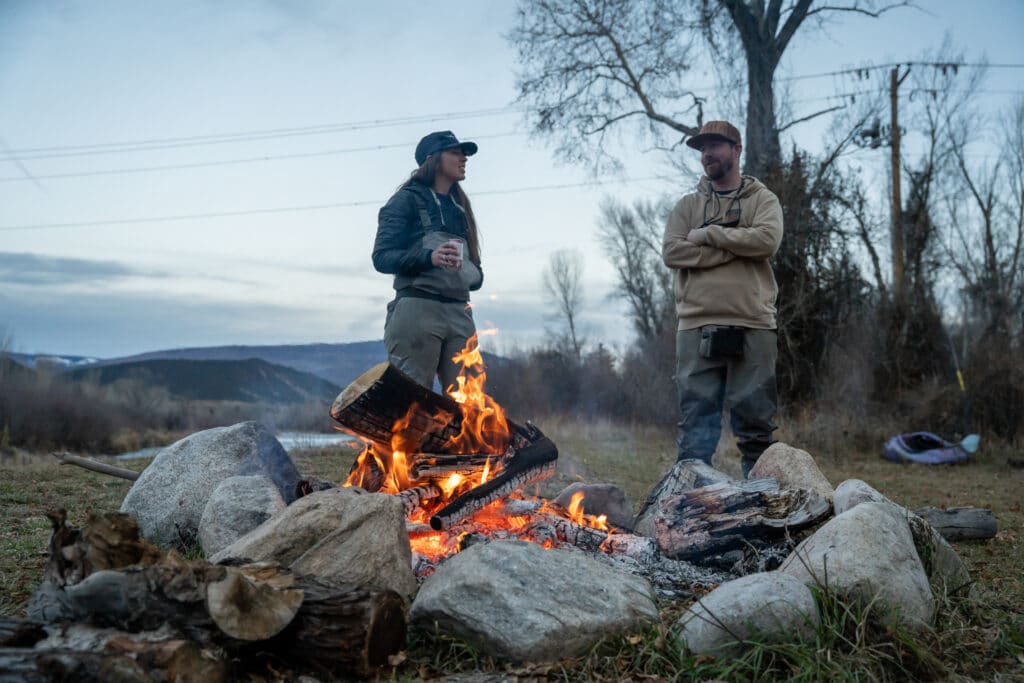 A man and a woman drink beer by a campfire beside a river in Colorado